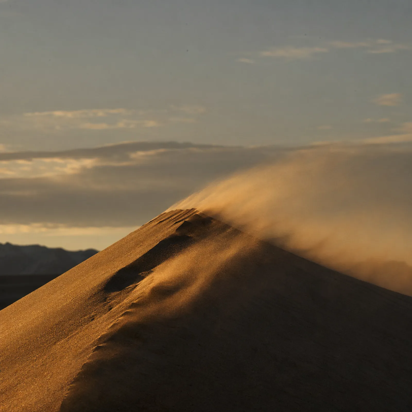 Wind lifting sand off the crest of a dune at sunset.